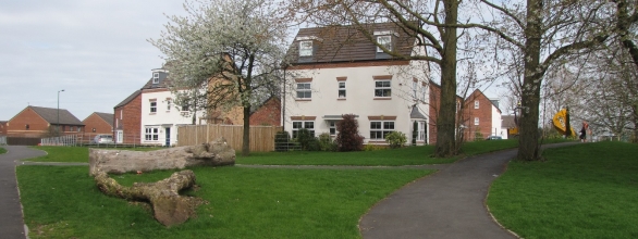 A white multi-storey building with several windows, surrounded by trees and a green lawn. A paved pathway curves through the grass, with a large fallen tree trunk in the foreground. Old and new residential buildings are visible in the background under an overcast sky.