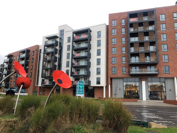 Three modern multi-storey apartment buildings with balconies, some with "sold" signs, behind a garden with tall grasses and red flower sculptures in the foreground.