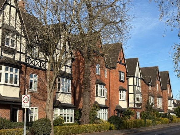 Three- and four-storey terraced houses with red brick and white and black timber framing, fronted by trees and shrubs, under a blue sky.