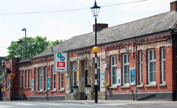 A red-brick building with multiple windows and a central entrance, located along a street with a lamppost and signage indicating a bus stop. There are posters on the walls and some bicycles parked nearby. Trees are visible in the background.