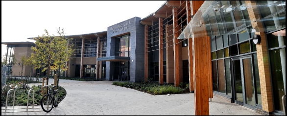 Modern building with curved facade, large glass windows, wooden vertical supports, and a covered entrance, surrounded by greenery and a paved pathway.