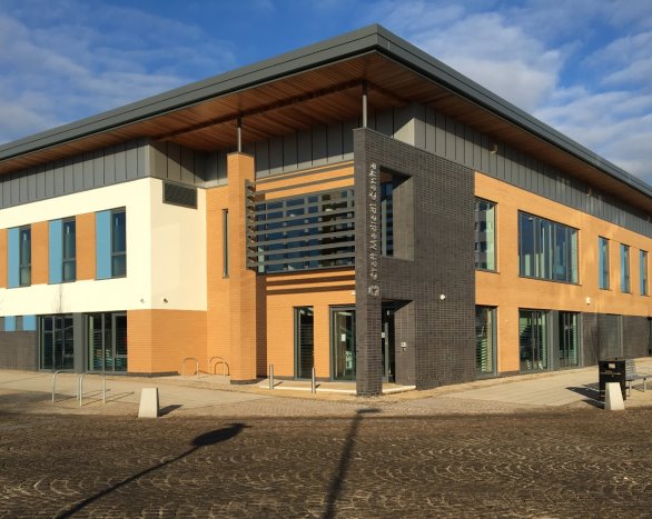 A modern two-storey building with a mix of beige, black, and orange brick exterior, large windows, and a flat roof. The entrance area features vertical black brick accents and a sign with text, set against a blue sky with scattered clouds.