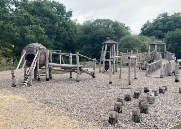 A wooden playground structure resembling a medieval castle, with towers, bridges, a tunnel, and climbing elements, set on a gravel surface surrounded by trees.