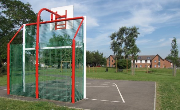 A basketball hoop attached to a fenced outdoor court with a surrounding grassy area, trees, and residential buildings in the background under a partly cloudy sky.
