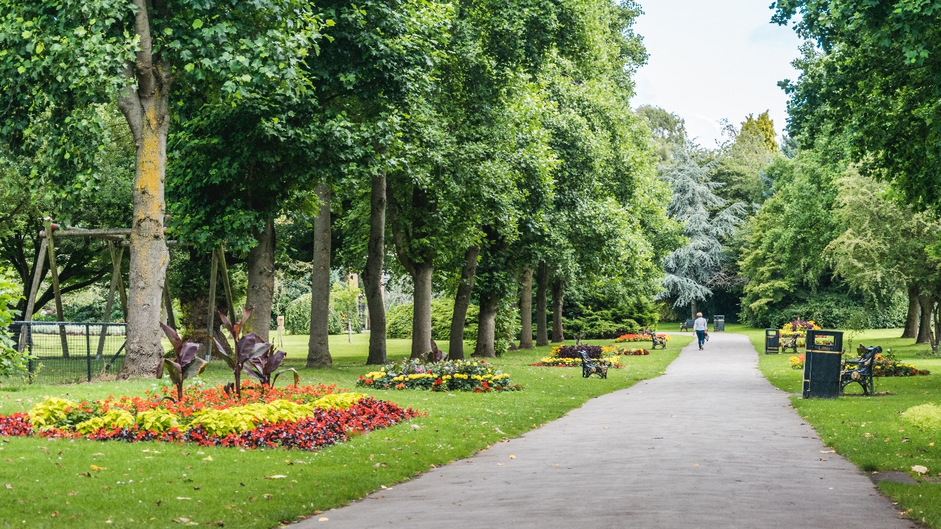 Photograph showing a landscaped public park with a paved pathway, mature trees, colourful flowerbeds and visitors. Formal planting, benches, bins and park signage line pathway, creating a peaceful recreational setting.