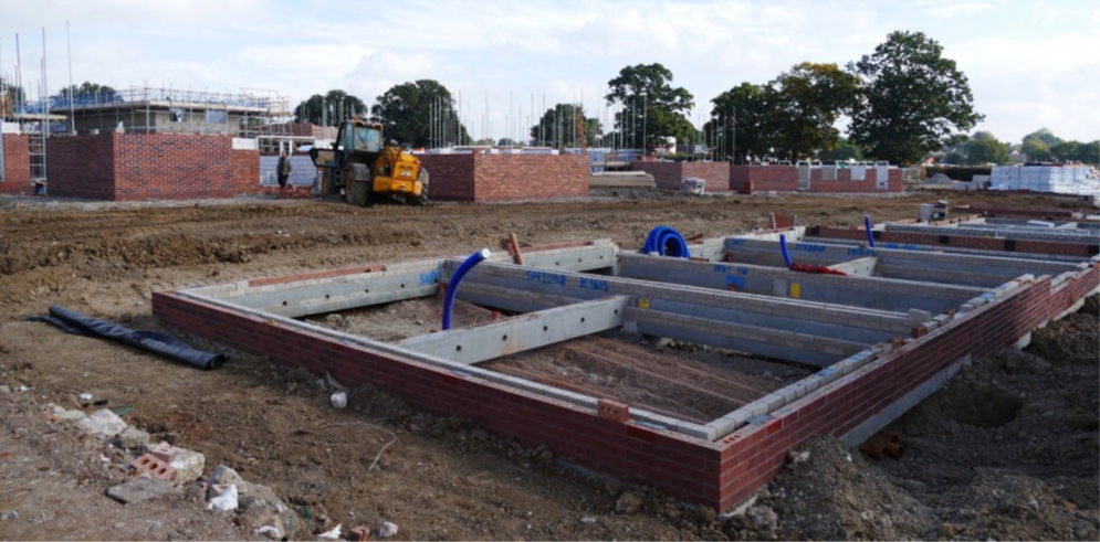 Photograph of a building site showing foundations of several houses and 4-5 rows of bricks. There is a digger and building materials as well as soil from the dug ground.
