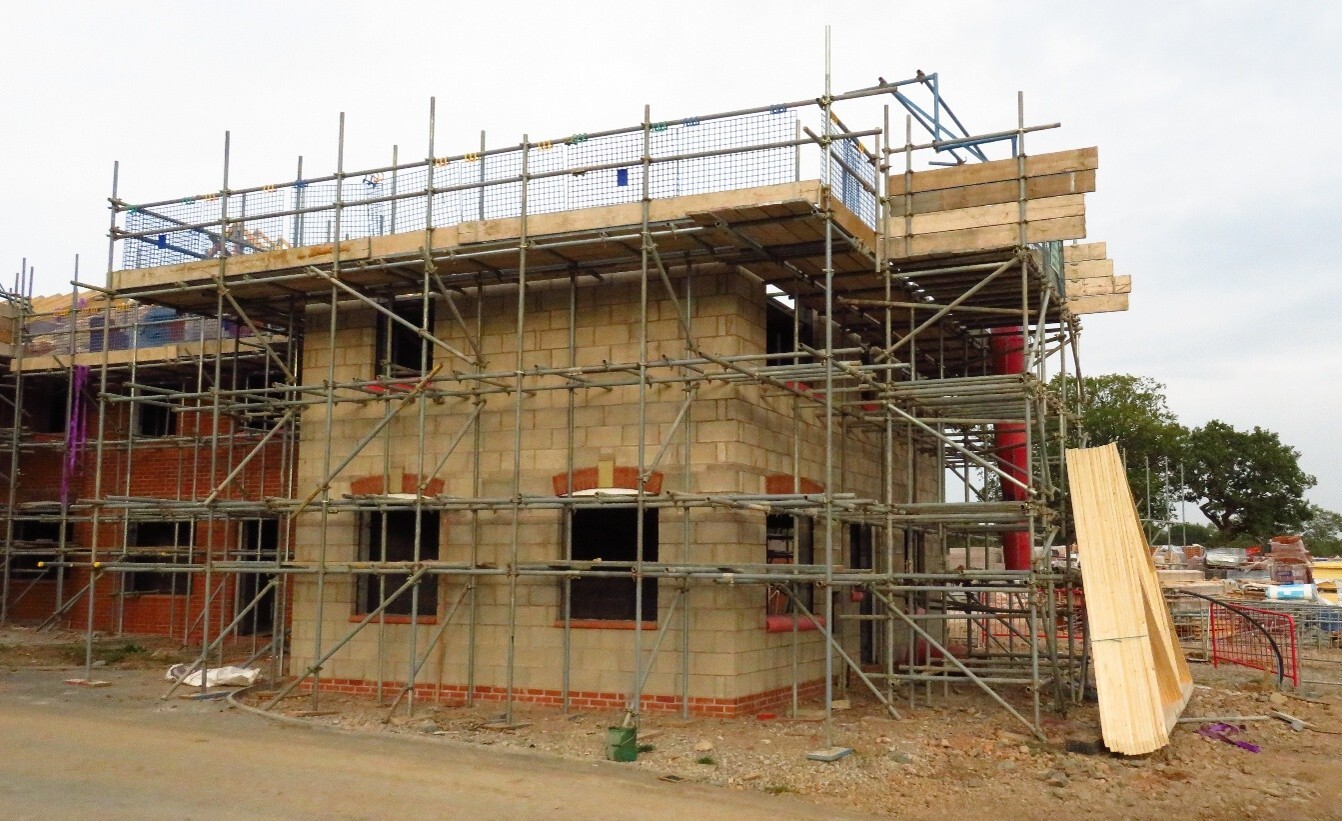 Photograph showing a two-storey house under construction, surrounded by metal scaffolding. Exposed blockwork, timber formwork, unfinished window openings, safety netting and stacked building materials highlight progress towards structural completion.
