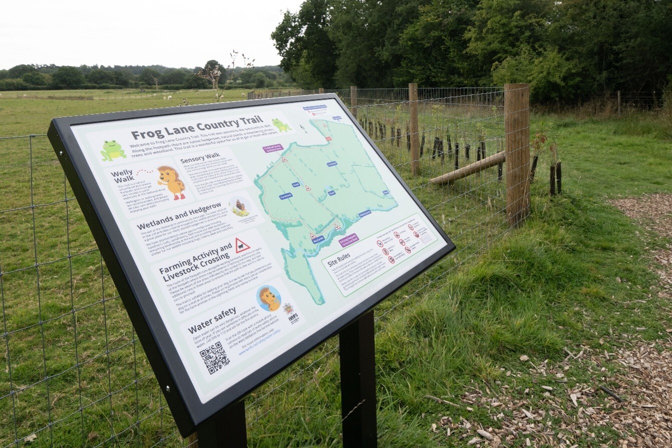 Photograph showing an outdoor information board beside a fenced rural footpath. The board is titled Frog Lane Country Trail and includes a coloured route map, wildlife and walking guidance, farming-safety warnings, water-safety advice, QR code and directional markers.