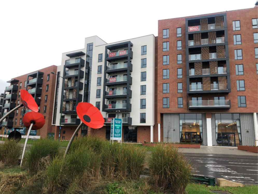 Photograph of medium rise modern apartment buildings with several 'SOLD' signs at front. The buildings are a mix of red brick, cream render with slate grey finishes on balconies. View taken from roundabout with large stylised poppies in foreground in natural vegetation.