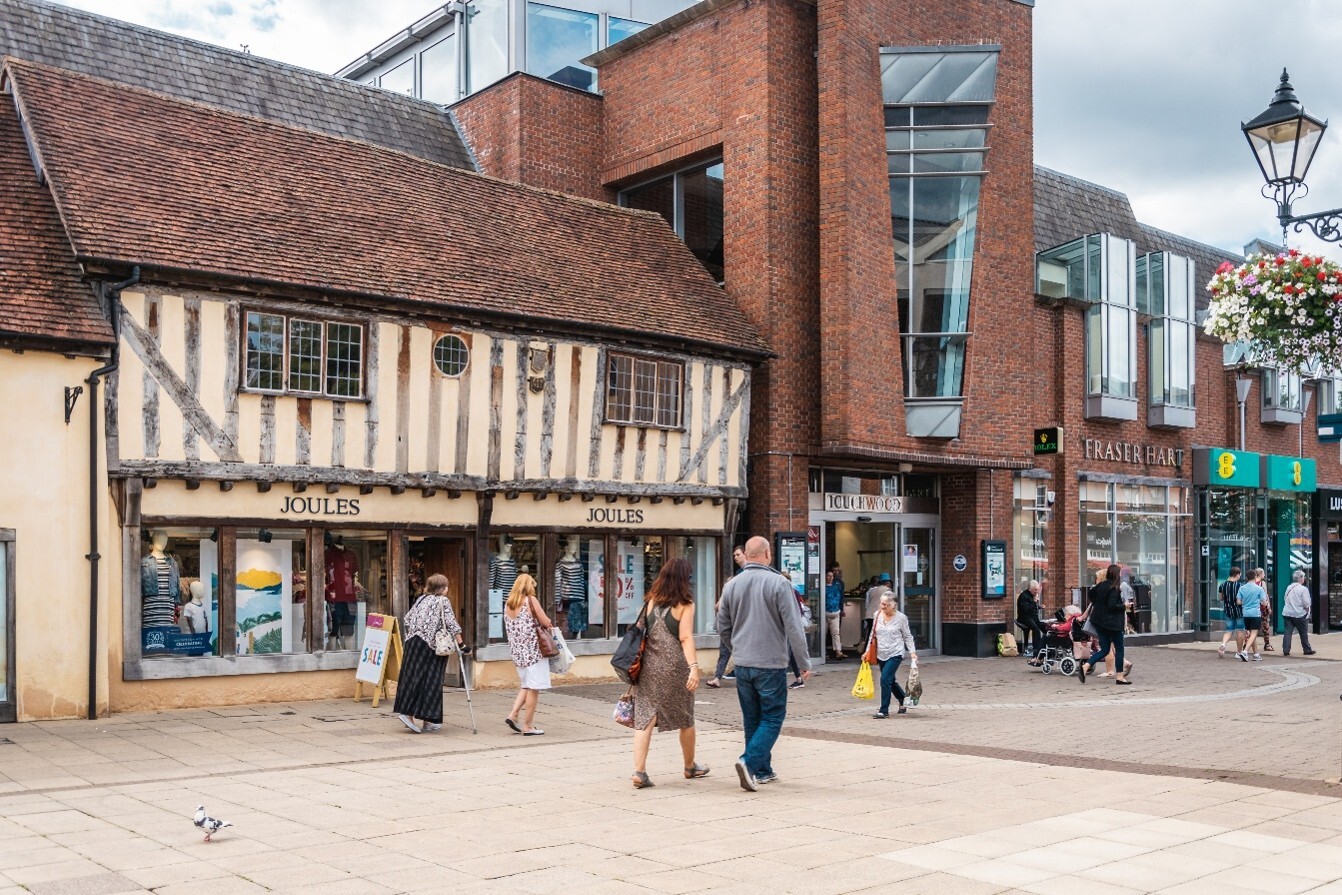 Photograph showing part of the High Street in Solihull Town Centre. The street is pedestrianised combining a timber-framed historic building with modern brick and glass architecture. Shoppers pass Joules, Fraser Hart and other shops, with hanging flower baskets, street lamps and colourful shopfront signs adding notable details.