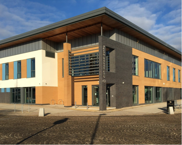 Photograph of large modern building with 'Croft Medical Centre' silver lettering on side. Building is flat roofed with mix of grey panel, brick and white render with large windows and glazed doors. Set next to hard landscaping with cycle rack, bench and public bin.