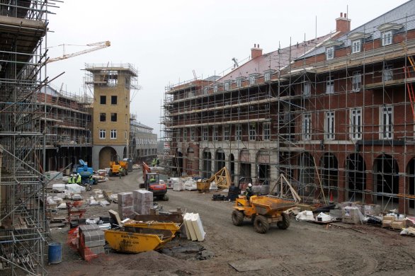 Photograph showing a construction site surrounded by scaffolded brick buildings and partially completed structures. The buildings are 4 to 5 storeys high with a mix of residential and commercial type buildings. Cranes, yellow dumper trucks, skips, pallets, building materials and unfinished façades are shown. 
