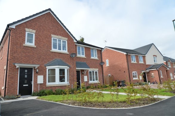 Photograph showing a newly built residential housing development with red-brick houses and landscaped front gardens. Two-storey homes feature dark tiled roofs, white-framed windows, bay windows, black doors and paved roads, with unfinished planting suggesting recent construction.
AI-generated content may be incorrect.