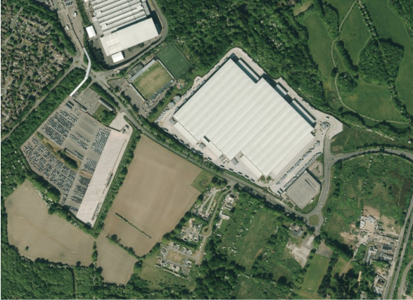 Aerial photograph showing a large industrial warehouse surrounded by access roads, farmland, woodland and residential areas. Nearby features include a substantial car park, sports pitches, smaller commercial buildings and a roundabout connecting local roads.