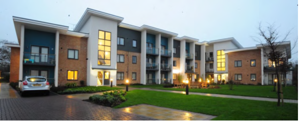 Photograph showing 2 to 3-storey apartment complex with mix of brick, dark panels and cream render facade. Some of the apartments have balconies with furniture. It is overcast with lights on bollards. The windows to the stairwells and of some apartments are illuminated. There is short mown grass landscaping at the front, block paving and a small tree. One car is on a driveway.