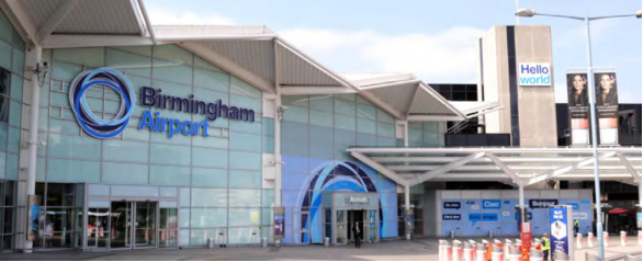 Photograph of main entrance to Birmingham Airport, with lettering shown in light and dark blue on glazed facade with large white awnings. There is a Hello World placard on a taller building and large advertisements with blurry picture of a woman's face in the background.