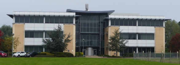 Photograph showing modern three-storey office building with symmetrical beige-and-glass frontage. Central glazed entrance, rooftop equipment, parked cars, landscaped grounds and overcast sky provide architectural and site context.
