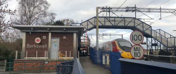 Photograph showing Berkswell railway station, with a passenger train approaching beside platform barriers and overhead electrification. Brick station building, footbridge, railway signals and circular speed restriction signs marked “100” and “MU 110” are visible.