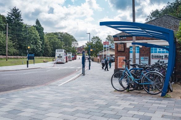 Photograph showing a public transport hub with a two-lane road, a zebra crossing and a blue cycle rack in the foreground, a bus stop with several buses on the left and the entrance to a railway station comprising a brick building on the right, and several pedestrians. There are mature trees, a landscaped verge, neat block paving with low rise buildings suggesting a suburban setting. The British Rail logo is mounted on tall white pillars. An “Evening Mail” advert and pavement markings add local street detail.