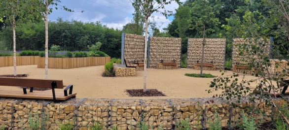 Photograph showing landscaped garden designed for relaxation and outdoor seating. Sandy paths, timber bench, young trees, gabion stone walls, planted borders and woven screening panels create a structured, natural setting.