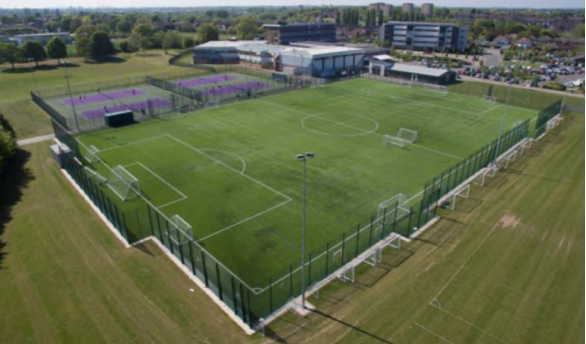 Aerial photograph of several playing pitches of different sizes on a sunny day with medium rise commercial buildings in the background and car parking.