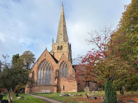 Photograph showing a historic brick St Alphege church with tall stone spire, Gothic arched stained-glass windows and surrounding graveyard. Autumn trees frame church, while gravestones, paved paths and manicured grounds suggest a tranquil resting place.