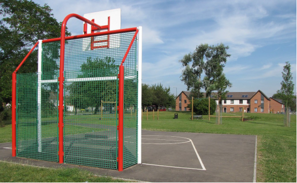 Photograph showing the back of a basketball hoop mounted on red metal pillars and green net surround with a small area of tarmac for shooting practice. It is within a recreation ground setting, with mown grassland, scattered trees and residential buildings to the rear.