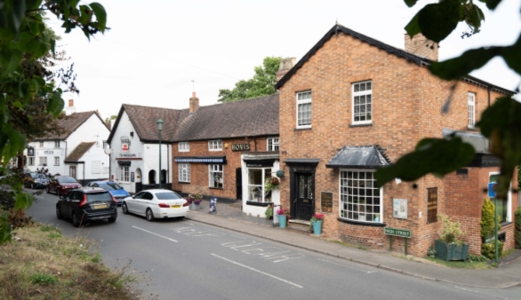 Photograph showing a high street lined with red-brick and white-painted buildings, small shops, parked cars and leafy trees. Street markings, shopfronts, hanging signs and residential architecture convey a village setting.