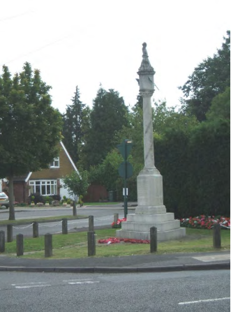 Photograph of war memorial which is a round column in stone set on a plinth. The memorial is set on grass with wreaths next to it and some flowers, and bordered with low wooden mounts and chain. It is next to a road with a house visible to the rear.