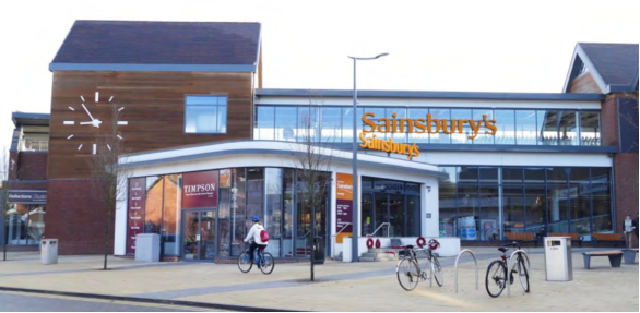 Photograph of a large Sainsbury's supermarket entrance, with orange lettering. Entrance surrounded by block paving with cycle stands, a bin, a cyclist and streetlamps. There is a large simplified clock face on the timber building facade.