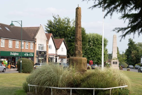 Photograph of village green in front of row of shops. There is a historic stone pillar in the centre of the green, a patch of ornamental and grass and shrubs and a pale stone obelisk to the rear of the green. The shops are a mix of red-brick and white render with tiled roof, including a 'Budgens'.