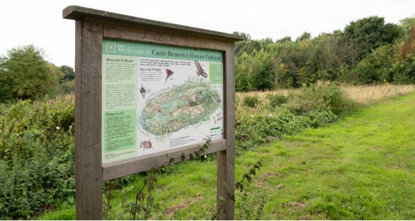 Photograph showing a wooden visitor information board welcoming visitors to Castle Bromwich Historic Parkland. Green panels display a site map, park history, walking routes and illustrated points of interest, set beside grassland and woodland.